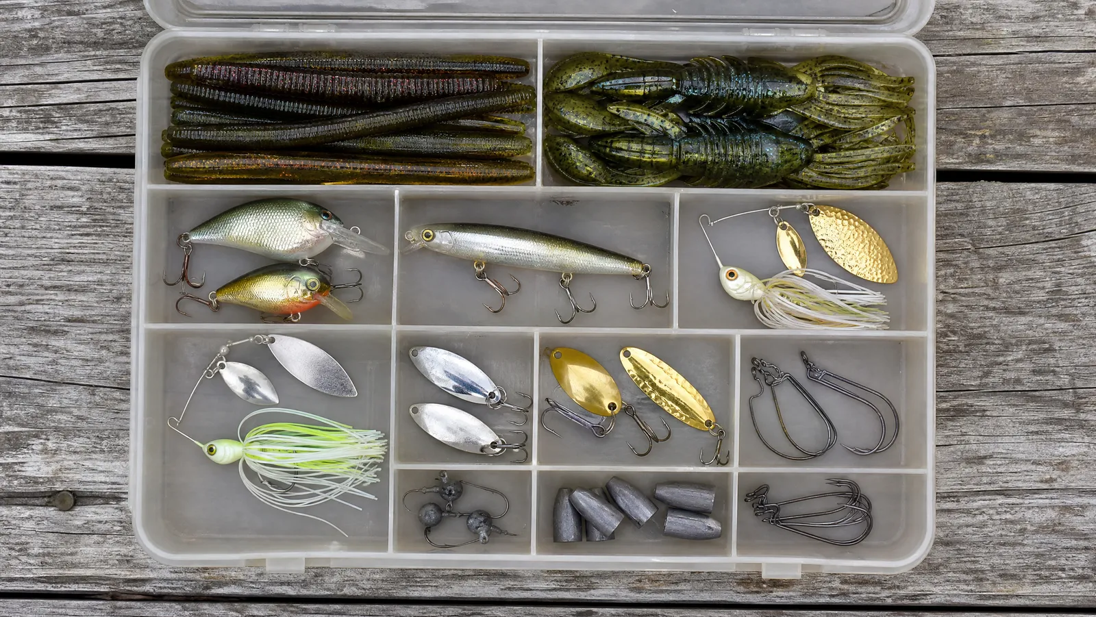 Open segmented tackle box on dock boards filled with soft plastics, crankbaits, spinners, spoons and hooks