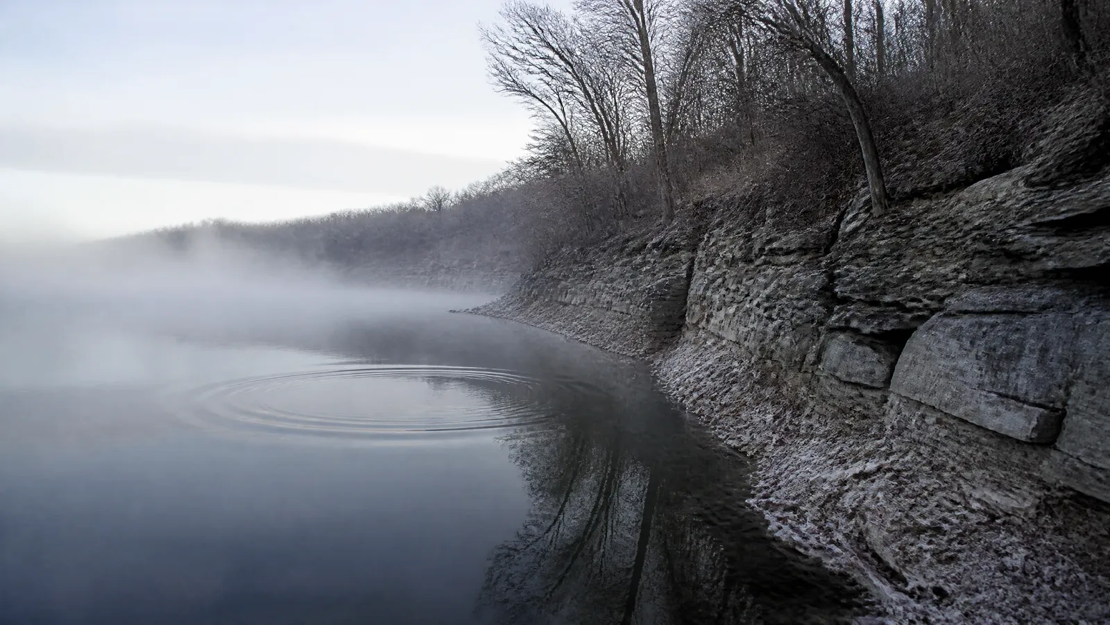 Steep grey rocky bluff bank dropping into slate-blue winter reservoir water under fog