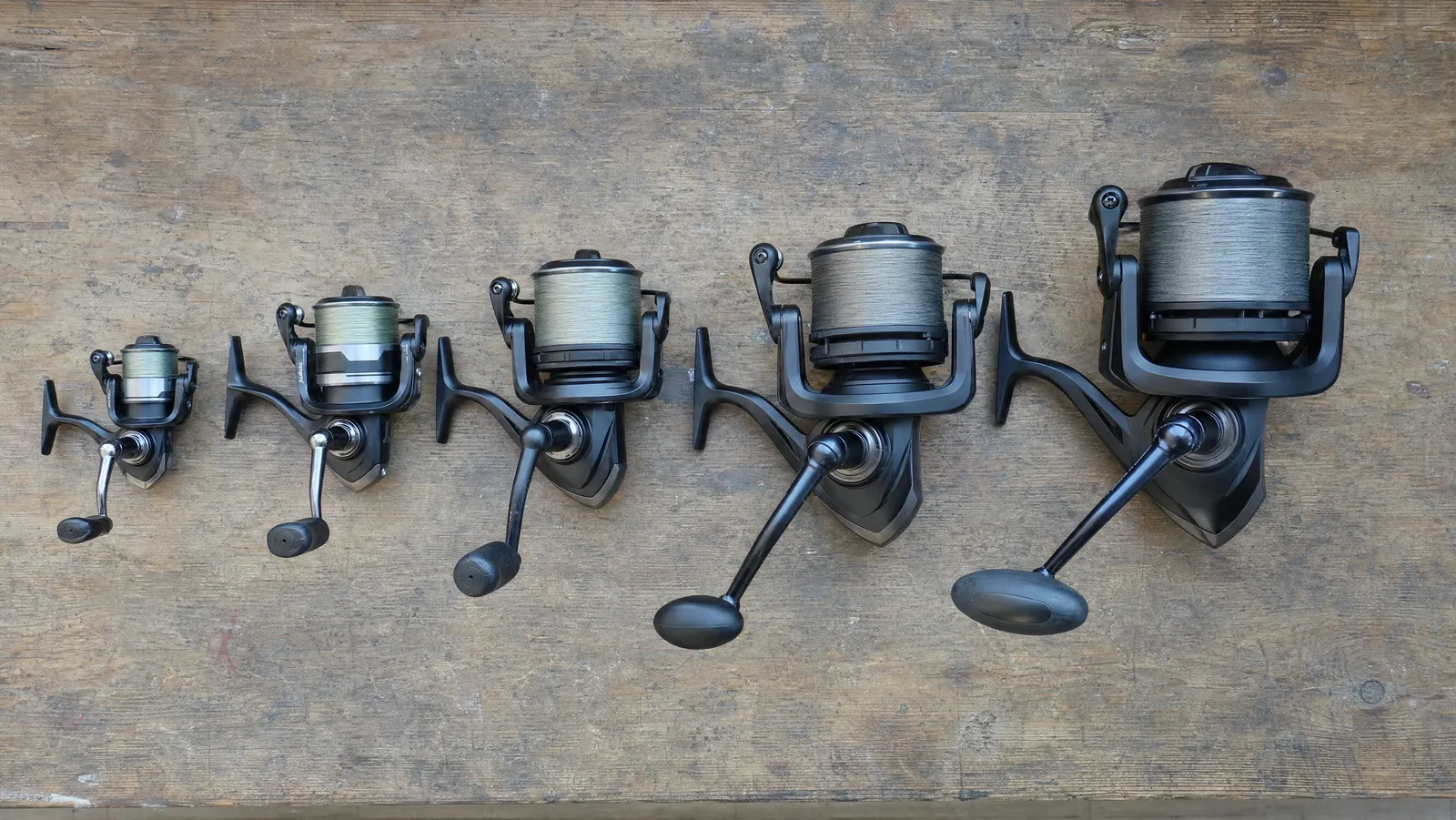 Five spinning reels laid out smallest to largest on a wooden workbench, showing how spool and body size grow through the size range