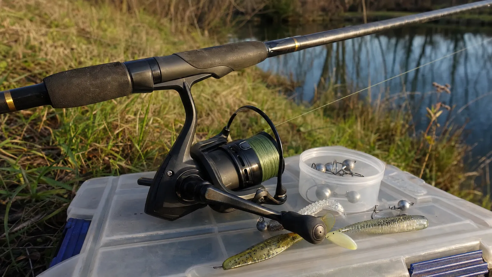 A matte black spinning reel mounted on a rod rests across a tackle box lid on a grassy pond bank in late afternoon light, with soft plastic lures beside it