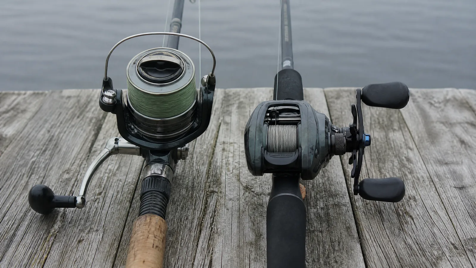 A spinning reel and a low-profile baitcasting reel laid side by side on weathered dock boards in flat morning light